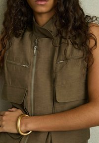 Khaki mouwloos vest met een hoge kraag, twee borstzakken en een voorkeursrits. Glad textuur met metalen accenten en een relaxed fit.