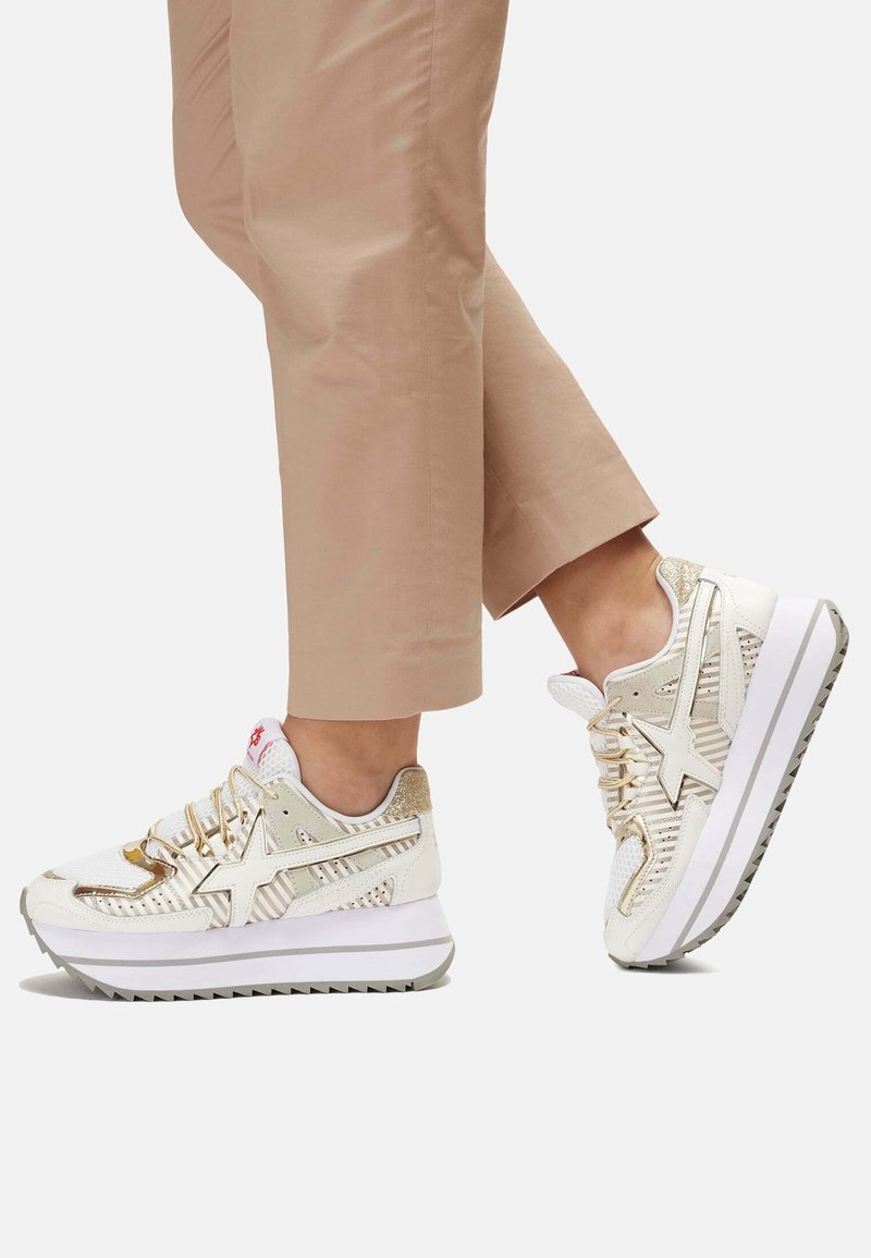 Witte en gouden sneaker met mesh en glanzende accenten, voorzien van een gestructureerde zool en veters. Geplaatst tegen beige broeken, die het ontwerp benadrukken.