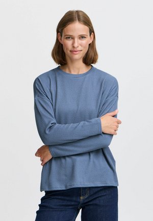 Lichtblauw, textuur gevend, lang mouwtop met een ronde halslijn en een losse pasvorm, gecombineerd met donkere jeans, met de armen gekruist voor de borst.