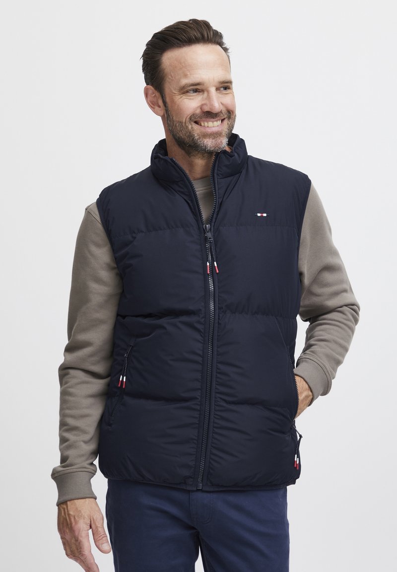 Mørkeblå puffer vest med høy krage, glidelås og sidelommer. Har diskrete logoaksenter og en kviltet design for ekstra tekstur.