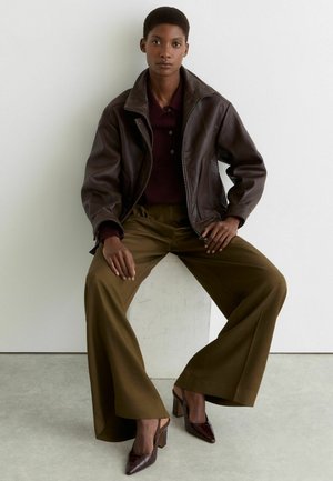 Woman sitting on a white block wearing a dark brown leather jacket, maroon shirt, olive wide-leg pants, and brown high-heeled shoes.