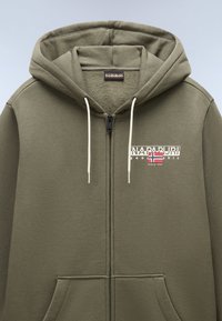 Felpa verde con zip e interno morbido, con lacci bianchi e un logo frontale che presenta una bandiera norvegese e il nome del marchio.