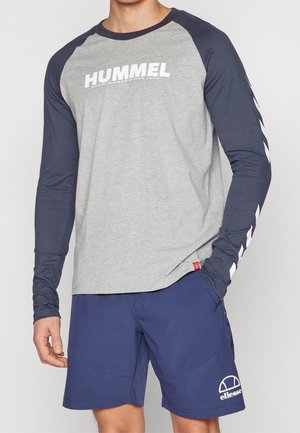 Mężczyzna w szarej i granatowej koszulce na długi rękaw marki Hummel oraz granatowych szortach Ellesse, stoi z jedną ręką w kieszeni na jednolitym tle.