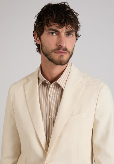 Man met kort krullend haar en baard, gekleed in een crèmekleurig colbert over een beige-wit gestreept overhemd, tegen een effen achtergrond.