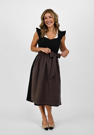 Smiling woman adjusts tied dark apron over black dress with ruffled sleeves, standing barefoot against plain light background.
