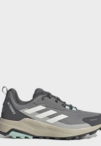 Chaussure de trail Adidas Terrex grise et noire avec des bandes blanches, semelle beige et semelle extérieure texturée pour les activités de plein air.