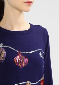 Jersey azul marino con adornos de lentejuelas coloridas, cuello redondo y una textura suave. Los elefantes muestran embellecimientos sobre la tela.