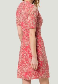 Lyhythihainen mekko korallinvärisenä kukkakuviolla. Mekossa on istuva siluetti ja rypytetty yksityiskohta vyötäröllä. Sujuva kankaan rakenne.