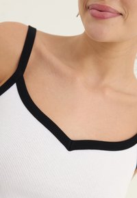 Top de tirantes blanco acanalado con un escote en V negro y tirantes. Textura suave con un ajuste ceñido, que resalta la zona de la clavícula.