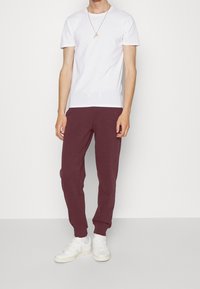 Person in einem weißen Kurzarmshirt, kastanienbraunen Jogginghosen, weißen Sneakers und einer goldenen Halskette, steht vor einem schlichten weißen Hintergrund.