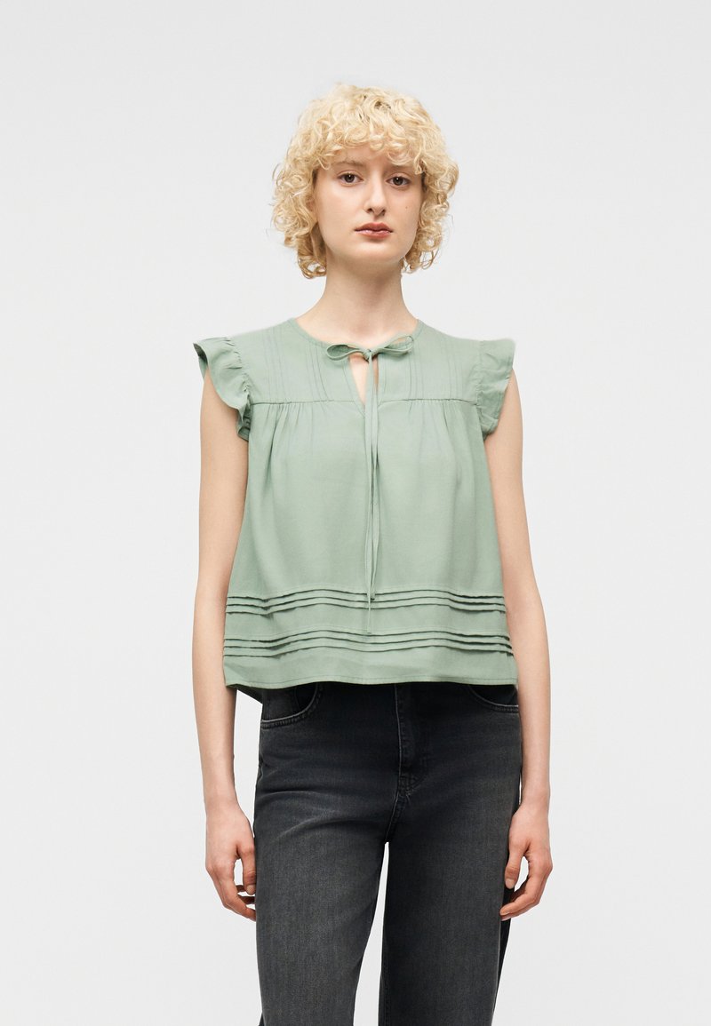 Νεαρός ενήλικας με σγουρά ξανθά μαλλιά, που φοράει μια αμάνικη μπλούζα σε μέντα πράσινο χρώμα με βολάν στους ώμους και σκούρο τζιν, στέκεται μπροστά σε απλό φόντο.