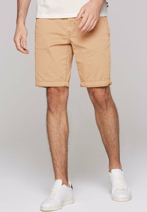 Man wearing beige rolled-hem shorts and white sneakers, standing against a neutral background.