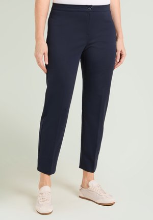 Donna che indossa pantaloni alla caviglia aderenti color blu navy e sneaker rosa pallido, in piedi davanti a uno sfondo neutro chiaro.