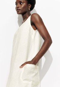 Robe sans manches de couleur claire avec un tissu texturé, présentant des motifs circulaires et des poches latérales, présentée sur un mannequin.