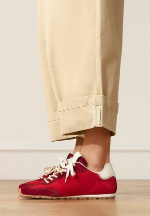 Persoon draagt beige cropped broek en rode suède sneakers met witte veters, staande op een houten vloer tegen een effen muur.