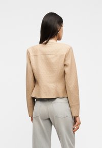 Γυναίκα με ίσια σκούρα μαλλιά που φοράει μπεζ σακάκι με σχέδια και ανοιχτόχρω μο γκρι τζιν, γυρισμένη με την πλάτη, σε απλό λευκό φόντο.