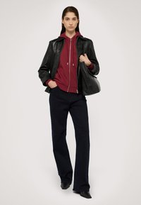Giacca di pelle nera sopra una felpa con cappuccio bordeaux, abbinata a jeans svasati blu scuro e scarpe nere a punta. Si porta una borsa nera.