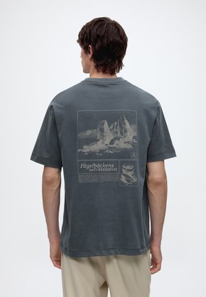 Mann trägt dunkelgraues T-Shirt mit Berg- und Vogelillustrationen und dem Text "Fågelbäckens Naturreservat" auf dem Rücken.