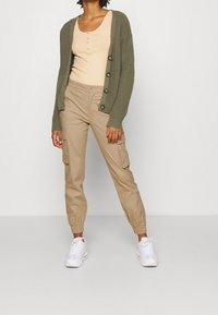 Cardigan côtelé vert sur un débardeur beige, pantalons cargo beiges avec des poignets élastiques, et baskets blanches. Comprend de gros boutons et des poches.