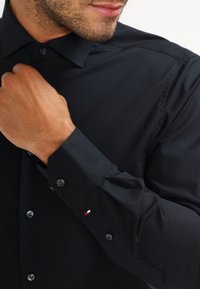 Camisa negra de manga larga hecha de tela suave, con cuello clásico, puños con botones y un pequeño detalle de logo tricolor en la manga.