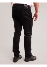 Férfi, aki fekete, szűk fazonú farmert és fekete tornacipőt visel, egyszerű világos háttér előtt áll, hátulnézetből.