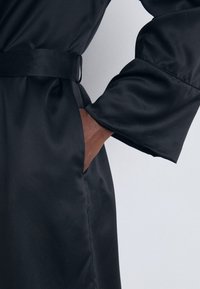 Svart satinrobe med bälte, med breda ärmar och en sidoficka. Tyget har en slät textur med en lätt lyster.