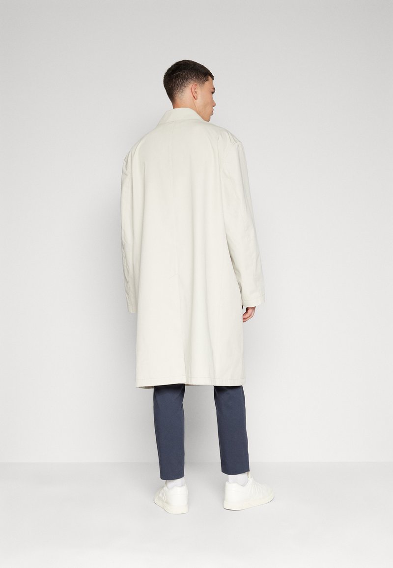 Cappotto trench grigio chiaro con vestibilità casual, design monopetto e spacco posteriore. Abbinato a pantaloni blu scuro e sneakers bianche.