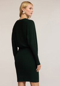 Abito lavorato a maglia verde scuro con maniche lunghe, silhouette aderente, scollatura rotonda e una texture liscia. La vista posteriore mostra dettagli delle cuciture e lunghezza fino al ginocchio.