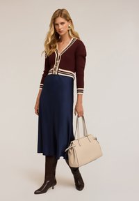 Cardigan tricotat în borș cu dungi crem, purtat împreună cu o fustă din satin albastru marin. Botine maro până la genunchi și o geantă bej completează ținuta.