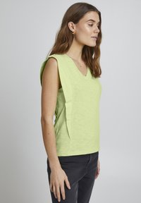 Top senza maniche verde chiaro con scollo a V, caratterizzato da un design con pannello laterale e una finitura leggermente textured. Indossato con pantaloni grigio scuro.