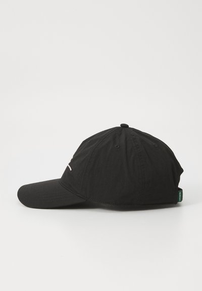 Casquette de baseball noire avec un visière incurvée, texture en tissu, et une petite étiquette verte sur le côté. Design simple avec des accents de couture.