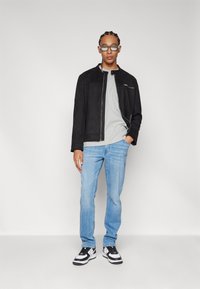 Chaqueta de ante negra con un cuello alto, camiseta gris debajo, jeans de color azul claro y zapatillas en negro y blanco. Gafas transparentes llevadas en el rostro.