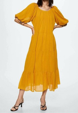 Kobieta w musztardowożółtej sukience midi z bufkami i falbaną na dole, do tego czarne sandały na obcasie z paskami.