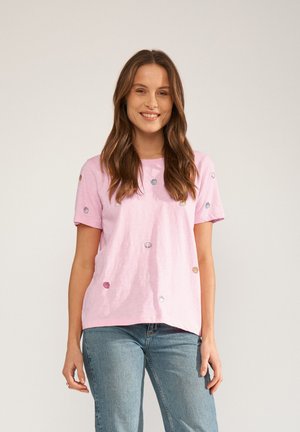 Femme aux longs cheveux bruns souriante, portant un t-shirt rose clair avec de petits motifs coquillages et un jean bleu, debout devant un fond uni.
