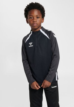 Dreng med kort krøllet hår og fregner, iført en sort og grå sportstop og sorte bukser med Hummel-logoer på en ensfarvet baggrund.