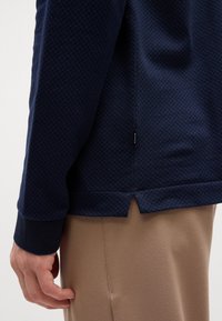 Pullover azul marinho com textura e caimento longo nas costas, com punhos em contraste, combinado com calças bege. Apresenta um pequeno rótulo na costura lateral.