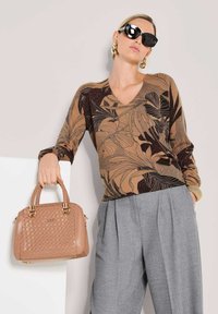 Vrouw in een geblokt bruine top en grijze broek, die een gestructureerde lichtbruine handtas met gevlochten handvatten vasthoudt, met zwarte zonnebril op.