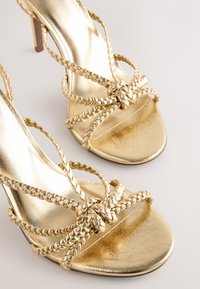 Sandales à talons hauts dorées avec des brides tressées et un design noué sur le dessus des orteils. La surface est brillante avec une texture lisse.