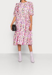 Květinové midi šaty v růžové a fialové barvě s nabíranými rukávy, zúženými patry a kulatým výstřihem. Nosené s levandulovými kotníkovými botami a černou kabelkou.