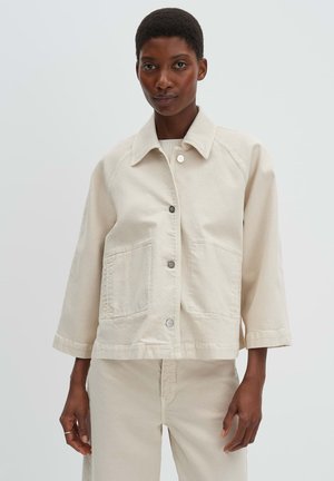 Frau trägt eine beige Jacke mit Knöpfen und großen vorderen Taschen sowie passende beige Hosen, steht vor einem einfachen hellen Hintergrund.