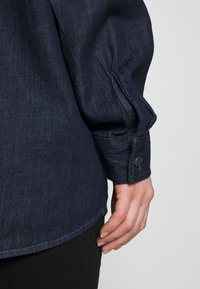 Gros plan d'une main reposant sur le côté, portant une chemise en denim bleu foncé à manches longues avec un poignet boutonné.