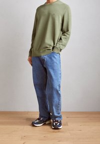 Mężczyzna w oliwkowozielonym swetrze z długim rękawem, luźnych niebieskich dżinsach i wielokolorowych trampkach stoi na jasnej drewnianej podłodze na tle gładkiej ściany.