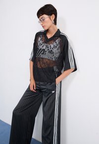 Black lace shirt with Adidas logo over a striped black and white track pant featuring side stripes, relaxed fit, and wide legs.