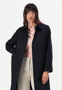 Marineblauer, taillierter Außenmantel mit klassischem Kragen, Knopfverschluss vorne und Seitentaschen. Über einem hellrosa Oberteil und hellen Hosen getragen.