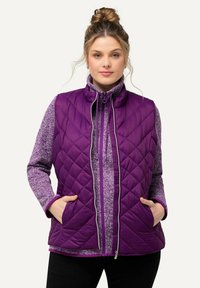Lila gesteppte Weste mit hohem Kragen, Reißverschluss vorne und zwei Taschen, über einem strukturierten lilafarbenen Pullover getragen, vor einem neutralen Hintergrund.