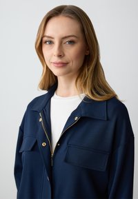 Giacca a zip blu navy in tessuto testurizzato con due tasche sul petto e dettagli in metallo dorato. Livello interno bianco visibile.