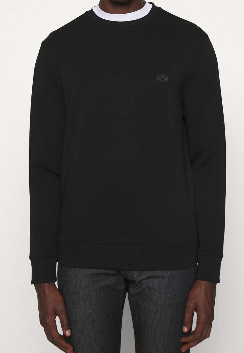 Man i en svart långärmad tröja med rund hals över en vit undertröja, tillsammans med mörkgrå byxor.