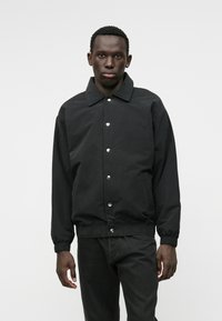 Melns bumbiera stila žakets ar apkakli, pogu aizdari, elastīgām aprocēm un jostu, ar gludu, matētu apdari.