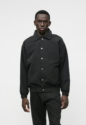 Veste bombardier noire avec un col, fermeture à boutons pression, poignets et taille élastiques, au fini lisse et mat.