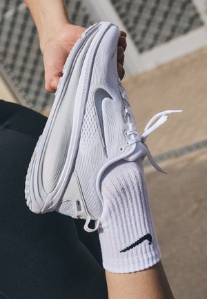 Hånd holder udstrakt ben iført hvid Nike-strømpe og hvid Nike-løbesko med sølv swoosh-logo udendørs.